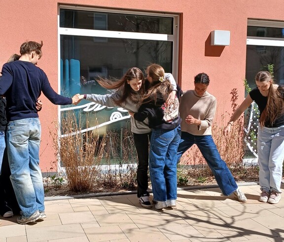 Schülerinnen und Schüler bei einem Teambuilding Spiel. Halten einander fest, stehen auf einem Blatt Papier zu zweit.