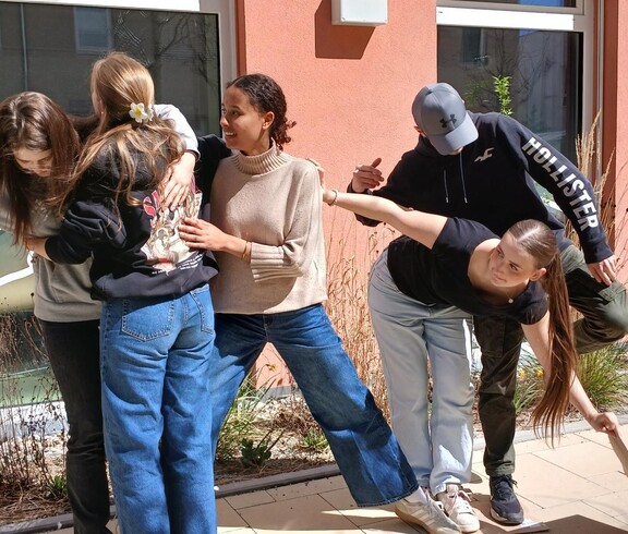 Schülerinnen und Schüler bei einem Teambuilding Spiel. Halten einander fest, stehen auf einem Blatt Papier zu zweit.