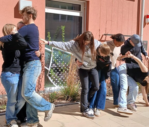 Schülerinnen und Schüler bei einem Teambuilding Spiel. Halten einander fest, stehen auf einem Blatt Papier zu zweit.
