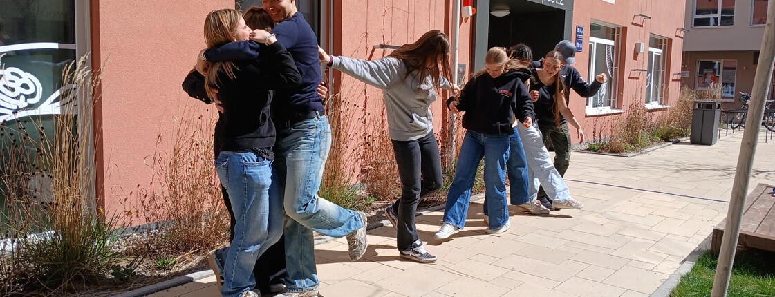 Schülerinnen und Schüler bei einem Teambuilding Spiel. Halten einander fest, stehen auf einem Blatt Papier zu zweit.