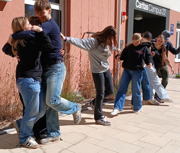 Schülerinnen und Schüler bei einem Teambuilding Spiel. Halten einander fest, stehen auf einem Blatt Papier zu zweit.